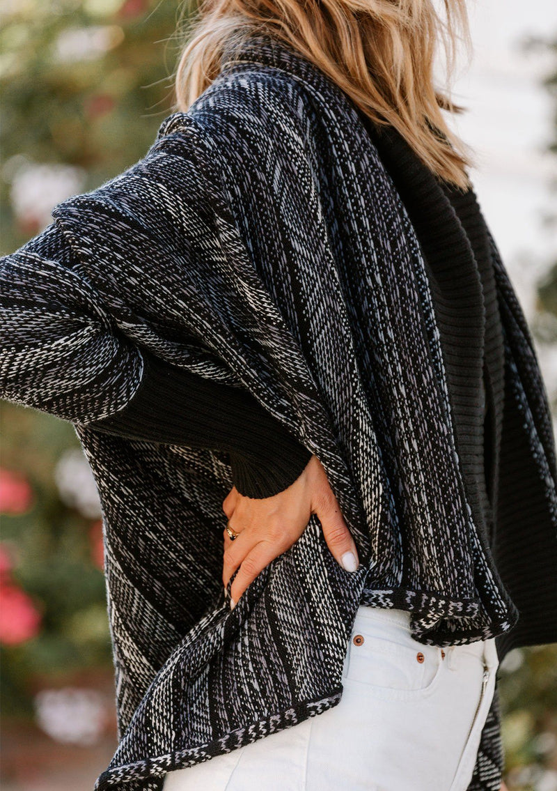 [Color: Black/Grey] A model wearing a cozy reversible shawl collar cardigan sweater in a black and grey southwestern inspired stripe jacquard. With long dolman sleeves and contrast ribbed trim. 