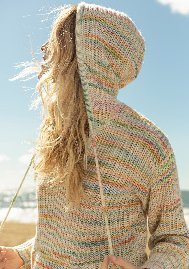 [Color: Sherbert] A close up side facing image of a blonde model at the beach wearing a multi color space dye knit sweater. A bohemian knit hoodie with long sleeves, a kangaroo front pocket, dropped shoulders, and a drawstring hood.