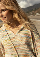 [Color: Sherbert] A close up front facing image of a blonde model at the beach wearing a multi color space dye knit sweater. A bohemian knit hoodie with long sleeves, a kangaroo front pocket, dropped shoulders, and a drawstring hood.