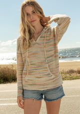 [Color: Sherbert] A front facing image of a blonde model at the beach wearing a multi color space dye knit sweater. A bohemian knit hoodie with long sleeves, a kangaroo front pocket, dropped shoulders, and a drawstring hood.