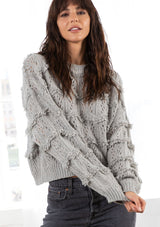 [Color: Grey] A model wearing a grey chunky knit sweater with long voluminous sleeves, a crew neckline, a cropped length and loop yarn fringe detail throughout. 
