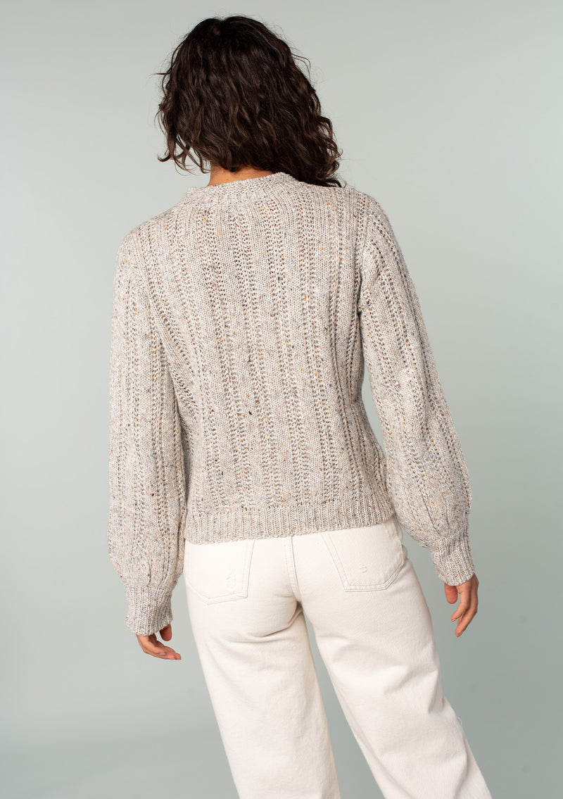 [Color: Oatmeal Multi] A back facing image of a brunette model wearing a perfect vintage style cream colored sweater with modern details. Voluminous long sleeves and a ladder stitch detail add a retro, yet timeless feel to this easy all season layer. Worn here with classic denim. 