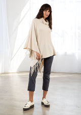 [Color: Vanilla] A model wearing a cream baby cable knit rib sweater poncho. With a cowl neckline and an asymmetric fringed hemline.