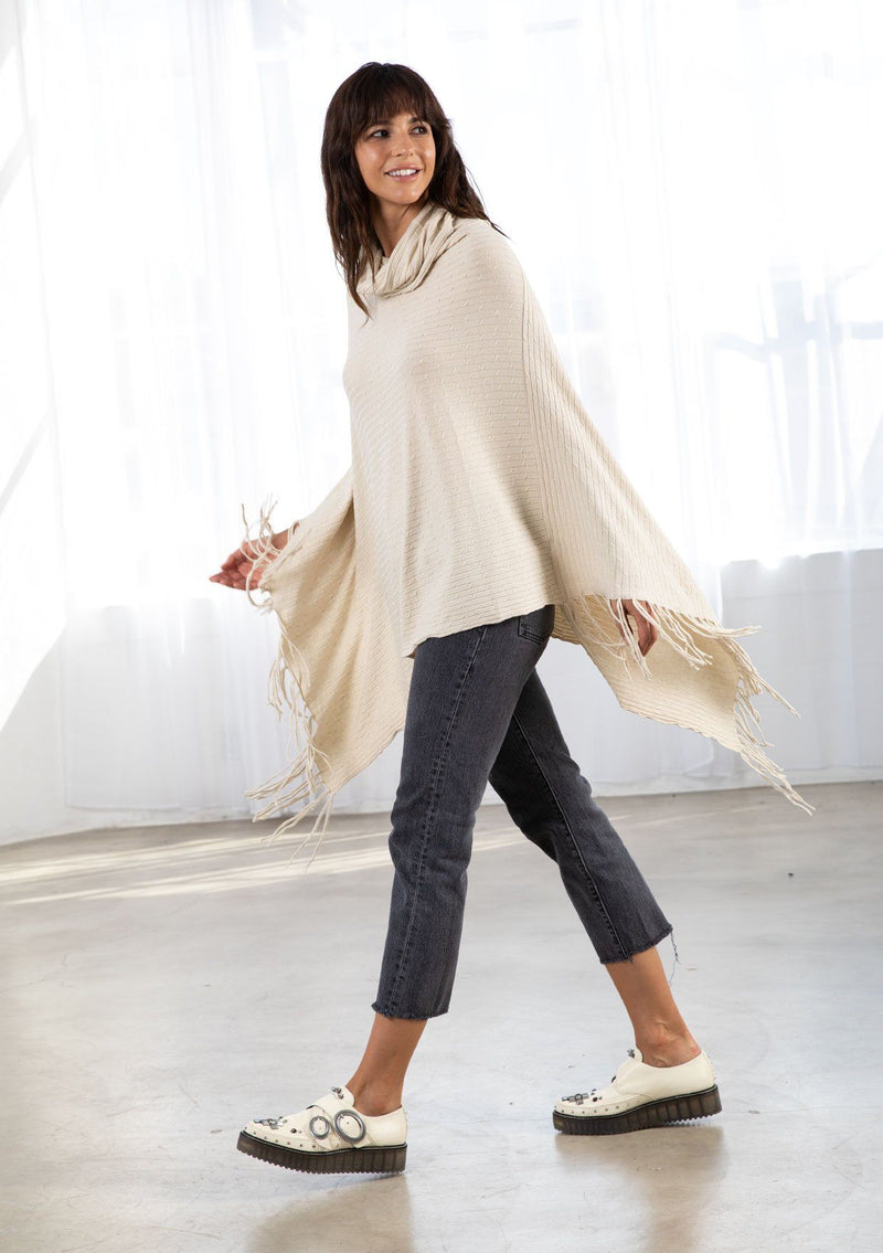[Color: Vanilla] A model wearing a cream baby cable knit rib sweater poncho. With a cowl neckline and an asymmetric fringed hemline.