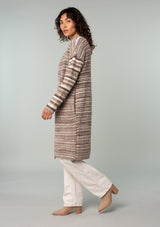 [Color: Brown/Taupe] A side facing image of a brunette model wearing a white and brown striped long cardigan. With an open front, contrast striped long sleeves, a shawl collar, and side pockets. 
