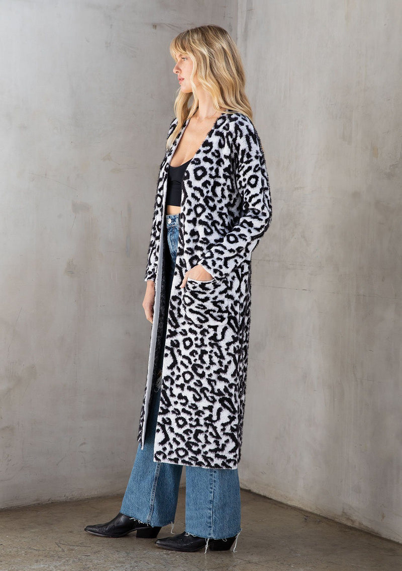 [Color: Snow Leopard] Ultra soft and fuzzy statement cardigan in a bold allover snow leopard print. A duster length sweater featuring oversize front patch pockets. Styled here with a black mini dress and boots.