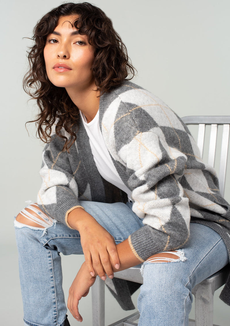 [Color: Charcoal/Grey] A sitting side facing image of a brunette model wearing an ultra soft mid length cardigan in a grey and light grey argyle plaid pattern. With long sleeves, an open front, and side pockets. 