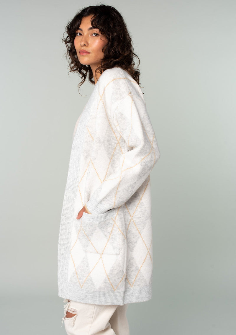 [Color: Heather Grey/Ivory] A side facing image of a brunette model wearing an ultra soft mid length cardigan in a light grey and ivory argyle plaid pattern. With long sleeves, an open front, and side pockets. 