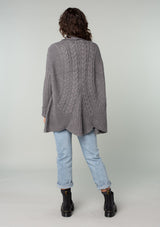 [Color: Heather Charcoal] A back facing image of a brunette model wearing a heather charcoal grey knit cardigan with a cocoon silhouette, braided cable knit detail, an open front, and a shawl collar. 