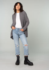 [Color: Heather Charcoal] A full body front facing image of a brunette model wearing a heather charcoal grey knit cardigan with a cocoon silhouette, braided cable knit detail, an open front, and a shawl collar. 