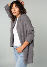 [Color: Heather Charcoal] A side facing image of a brunette model wearing a heather charcoal grey knit cardigan with a cocoon silhouette, braided cable knit detail, an open front, and a shawl collar. 