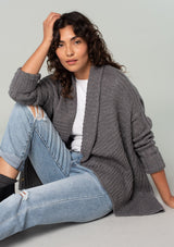 [Color: Heather Charcoal] A sitting side facing image of a brunette model wearing a heather charcoal grey knit cardigan with a cocoon silhouette, braided cable knit detail, an open front, and a shawl collar. 
