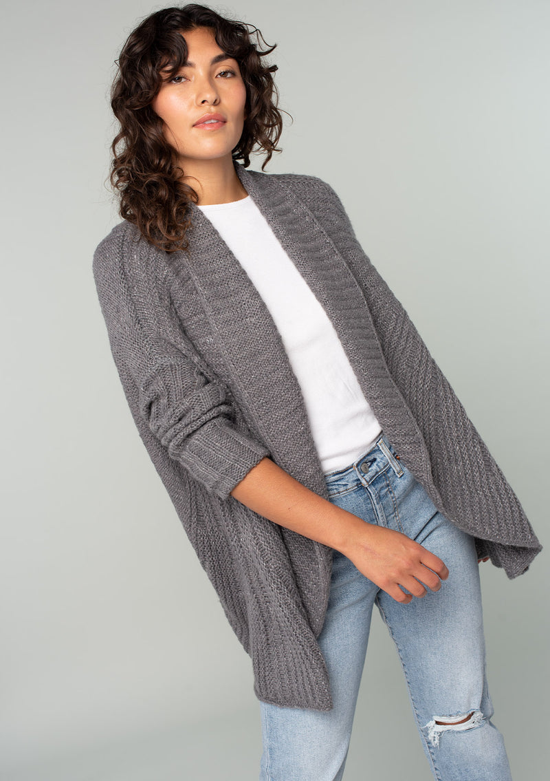 [Color: Heather Charcoal] A front facing image of a brunette model wearing a heather charcoal grey knit cardigan with a cocoon silhouette, braided cable knit detail, an open front, and a shawl collar. 