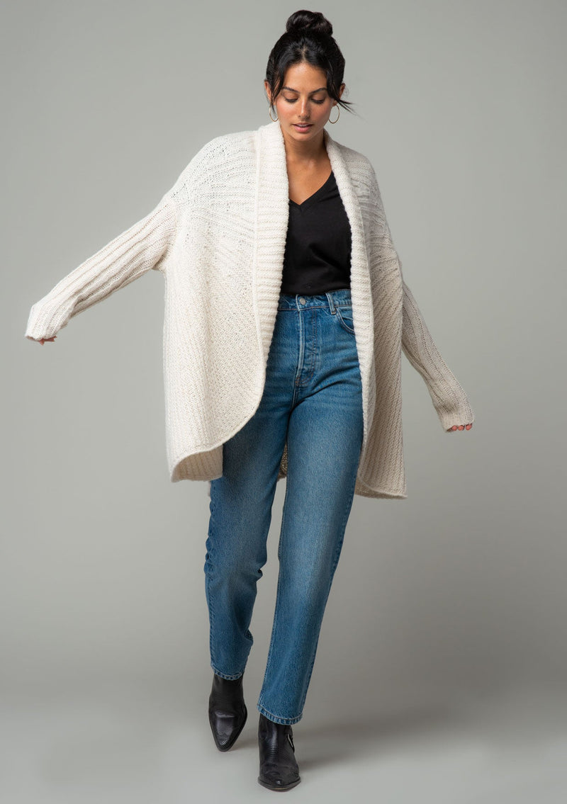 [Color: Oatmeal] A front facing image of a brunette model wearing a light oatmeal knit cardigan with a cocoon silhouette, braided cable knit detail, an open front, and a shawl collar. 