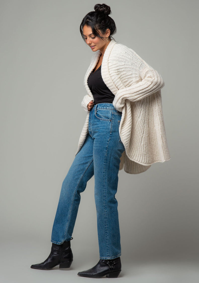 [Color: Oatmeal] A side facing image of a brunette model wearing a light oatmeal knit cardigan with a cocoon silhouette, braided cable knit detail, an open front, and a shawl collar. 