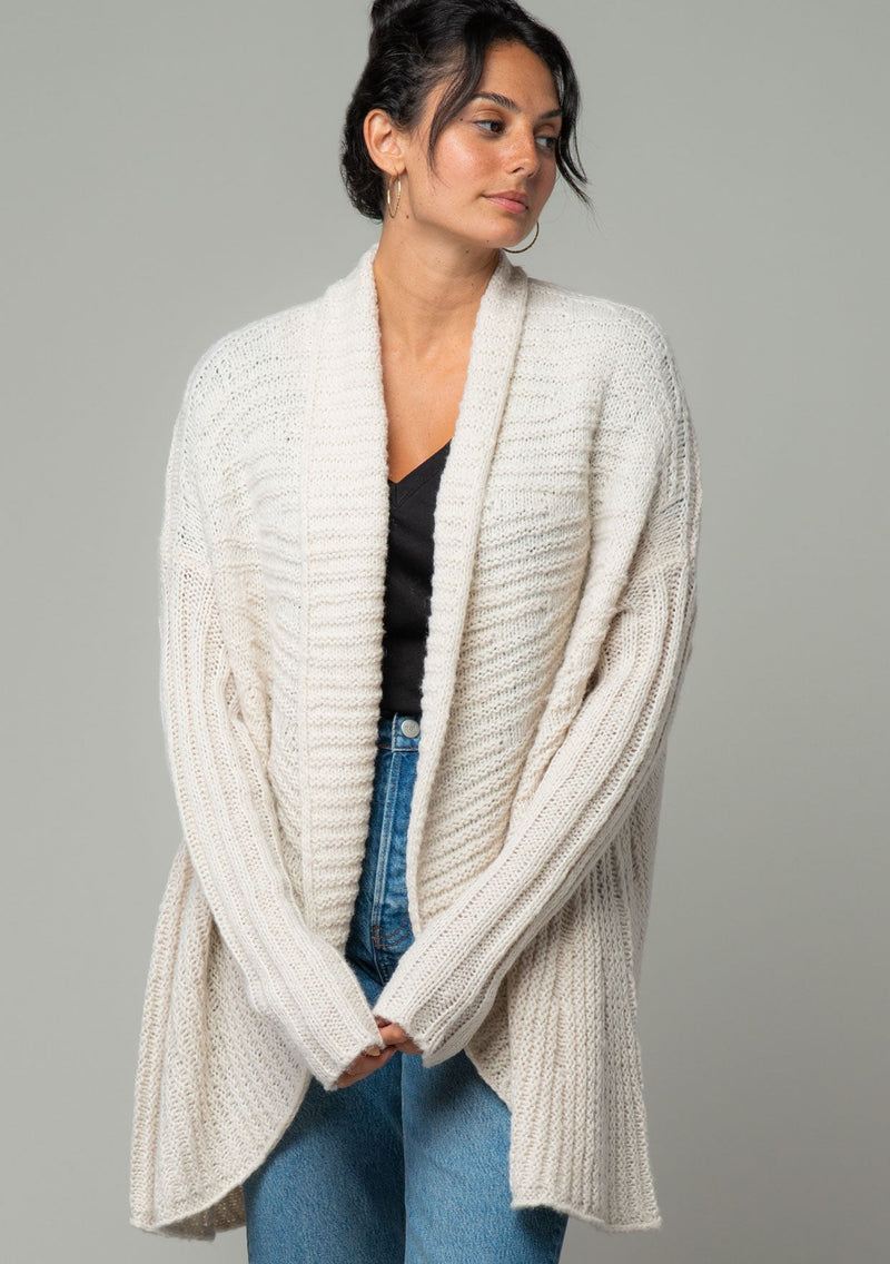 [Color: Oatmeal] A half body front facing image of a brunette model wearing a light oatmeal knit cardigan with a cocoon silhouette, braided cable knit detail, an open front, and a shawl collar. 