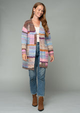 [Color: Pink Multi] A front facing image of a red headed model wearing a multi color pink patchwork knit cardigan. A mid length bohemian cardigan with long sleeves, an open front, and exposed seam details. 