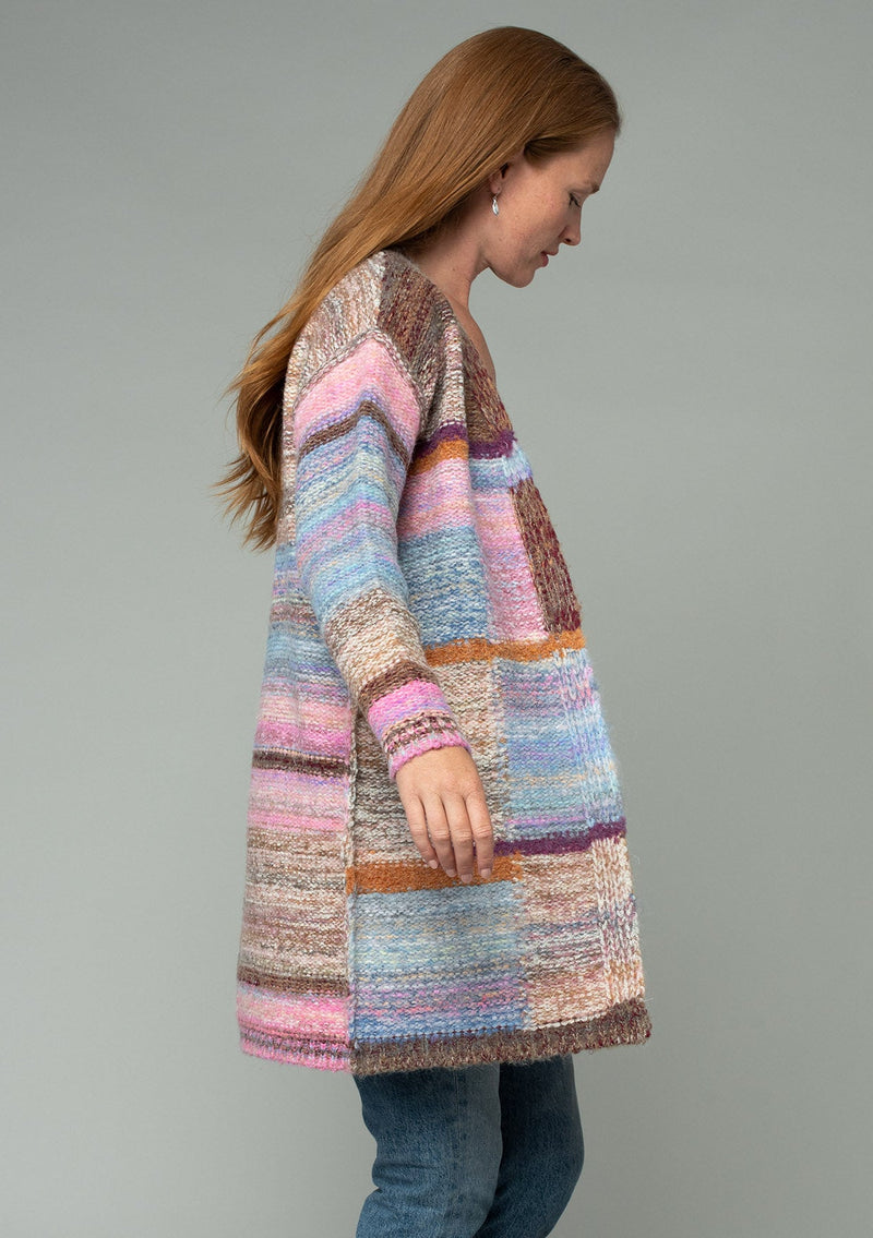 [Color: Pink Multi] A half body side facing image of a red headed model wearing a multi color pink patchwork knit cardigan. A mid length bohemian cardigan with long sleeves, an open front, and exposed seam details. 