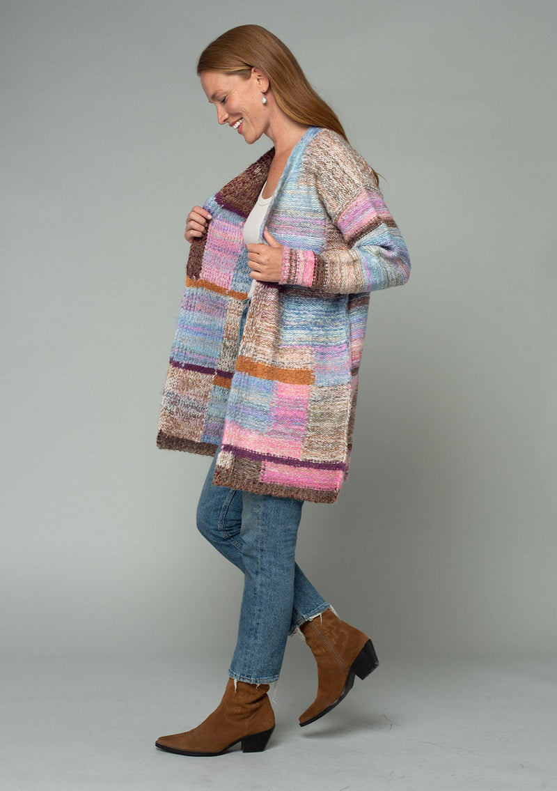 [Color: Pink Multi] A full body side facing image of a red headed model wearing a multi color pink patchwork knit cardigan. A mid length bohemian cardigan with long sleeves, an open front, and exposed seam details. 