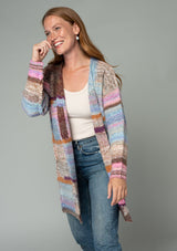 [Color: Pink Multi] A half body front facing image of a red headed model wearing a multi color pink patchwork knit cardigan. A mid length bohemian cardigan with long sleeves, an open front, and exposed seam details. 