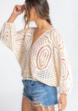 [Color: Natural] A model wearing a natural colored open knit bohemian crochet top with long voluminous sleeves and a wrap front. 