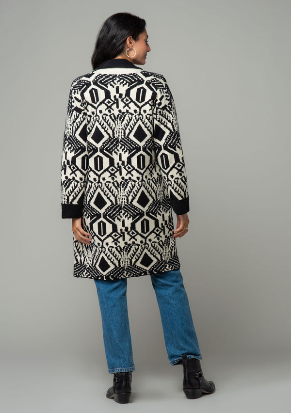 [Color: Natural/Black] A back facing image of a brunette model wearing a natural off white and black bohemian patterned mid length cardigan. A long sweater style with long sleeves, contrast black wrist cuffs, a contrast black shawl collar, an open front, and side pockets. 