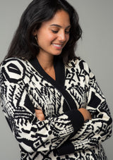 [Color: Natural/Black] A close up front facing image of a brunette model wearing a natural off white and black bohemian patterned mid length cardigan. A long sweater style with long sleeves, contrast black wrist cuffs, a contrast black shawl collar, an open front, and side pockets. 