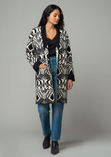 [Color: Natural/Black] A front facing image of a brunette model wearing a natural off white and black bohemian patterned mid length cardigan. A long sweater style with long sleeves, contrast black wrist cuffs, a contrast black shawl collar, an open front, and side pockets. 