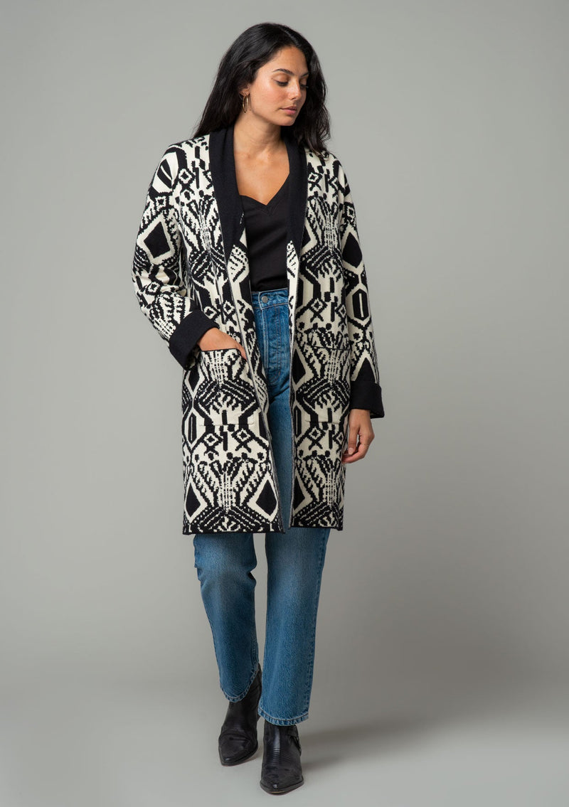 [Color: Natural/Black] A front facing image of a brunette model wearing a natural off white and black bohemian patterned mid length cardigan. A long sweater style with long sleeves, contrast black wrist cuffs, a contrast black shawl collar, an open front, and side pockets. 