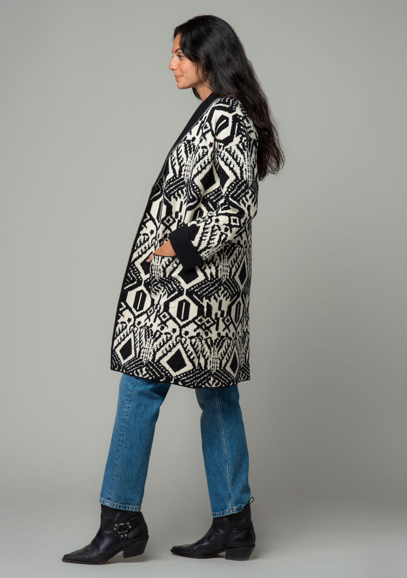 [Color: Natural/Black] A side facing image of a brunette model wearing a natural off white and black bohemian patterned mid length cardigan. A long sweater style with long sleeves, contrast black wrist cuffs, a contrast black shawl collar, an open front, and side pockets. 