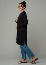 [Color: Black] A side facing image of a brunette model wearing a black waffle knit cardigan. A lightweight mid length cardigan sweater with long sleeves, side pockets, and an open front. 