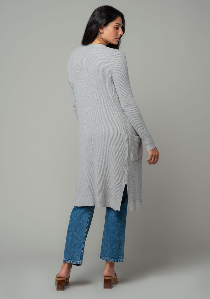 [Color: Heather Silver Grey] A back facing image of a brunette model wearing a light heather grey waffle knit cardigan. A lightweight mid length cardigan sweater with long sleeves, side pockets, and an open front. 