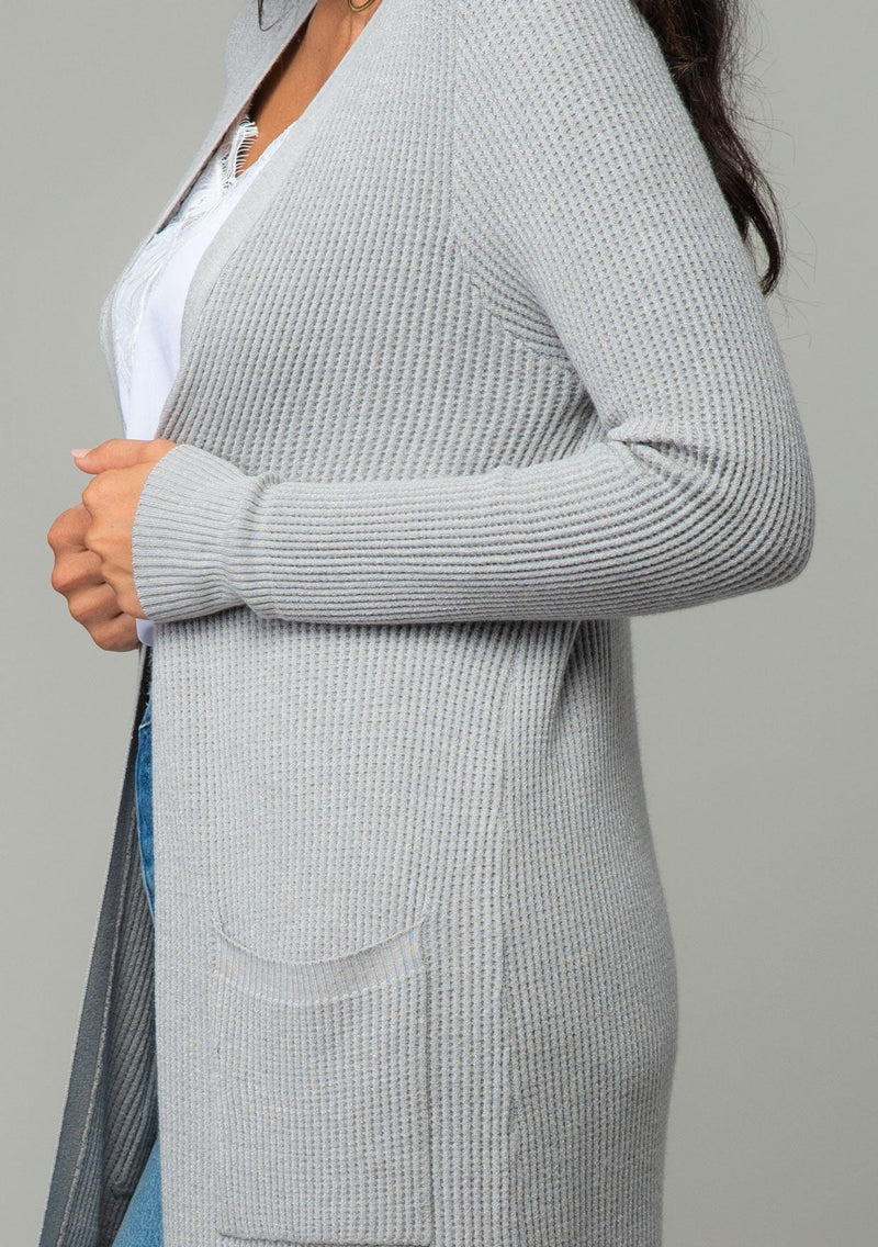 [Color: Heather Silver Grey] A close up side facing image of a brunette model wearing a light heather grey waffle knit cardigan. A lightweight mid length cardigan sweater with long sleeves, side pockets, and an open front. 