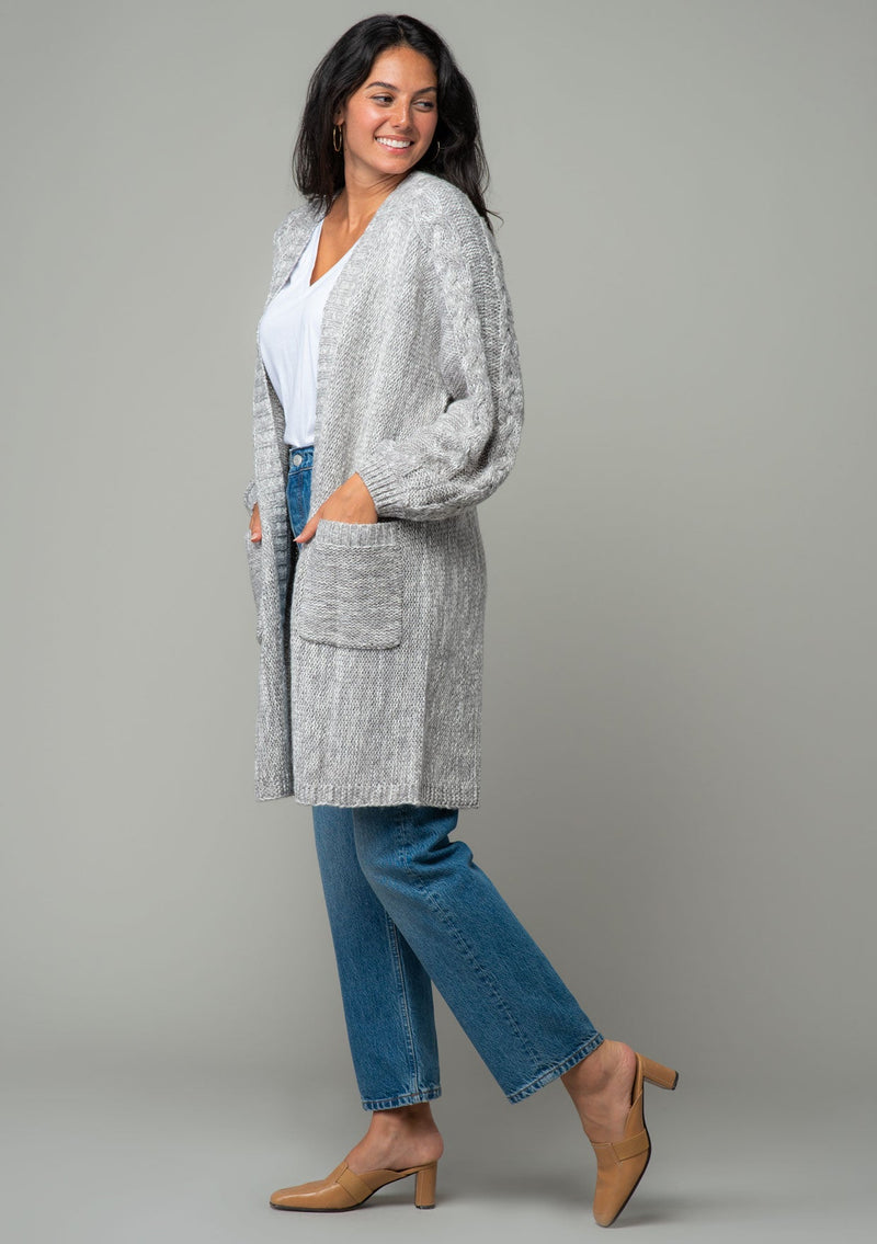 [Color: Heather Grey] A side facing image of a brunette model wearing a heather grey chunky knit cardigan. A fall sweater with long sleeves, cable knit sleeve detail, an open front, and side pockets. 