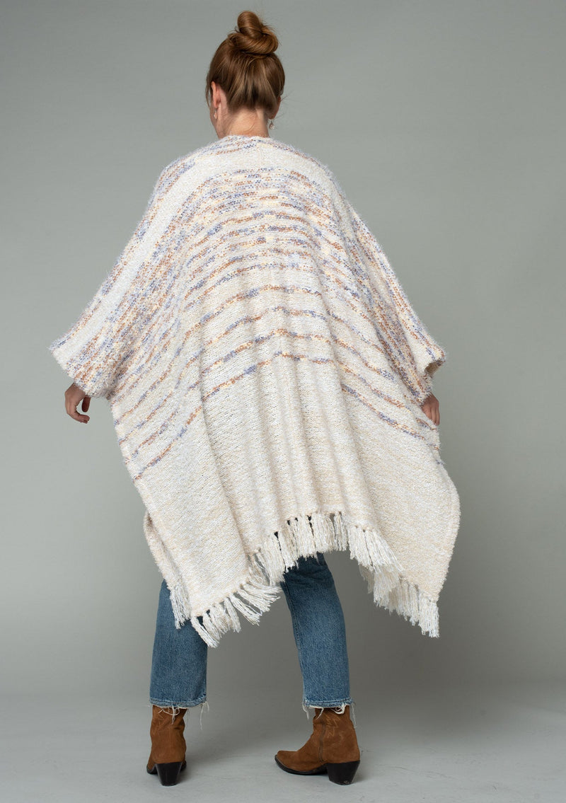 [Color: Oatmeal Multi] A back facing image of a red headed model wearing a fuzzy bohemian sweater poncho in a cream multi color knit. With an open front and tassel fringe trim. 