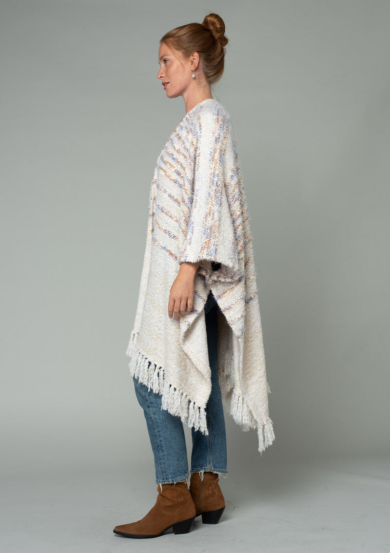 [Color: Oatmeal Multi] A side facing image of a red headed model wearing a fuzzy bohemian sweater poncho in a cream multi color knit. With an open front and tassel fringe trim. 