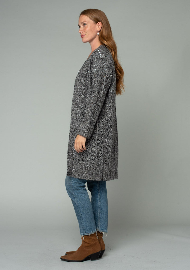 [Color: Charcoal] A side facing image of a red headed model wearing a dark charcoal grey open knit cardigan sweater. A mid length lightweight cardigan with long sleeves, an open front, and ribbed wrist cuffs. 