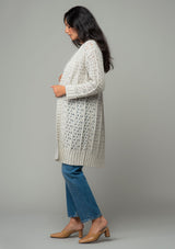 [Color: Oatmeal] A side facing image of a brunette model wearing an oatmeal open knit cardigan sweater. A mid length lightweight cardigan with long sleeves, an open front, and ribbed wrist cuffs. 