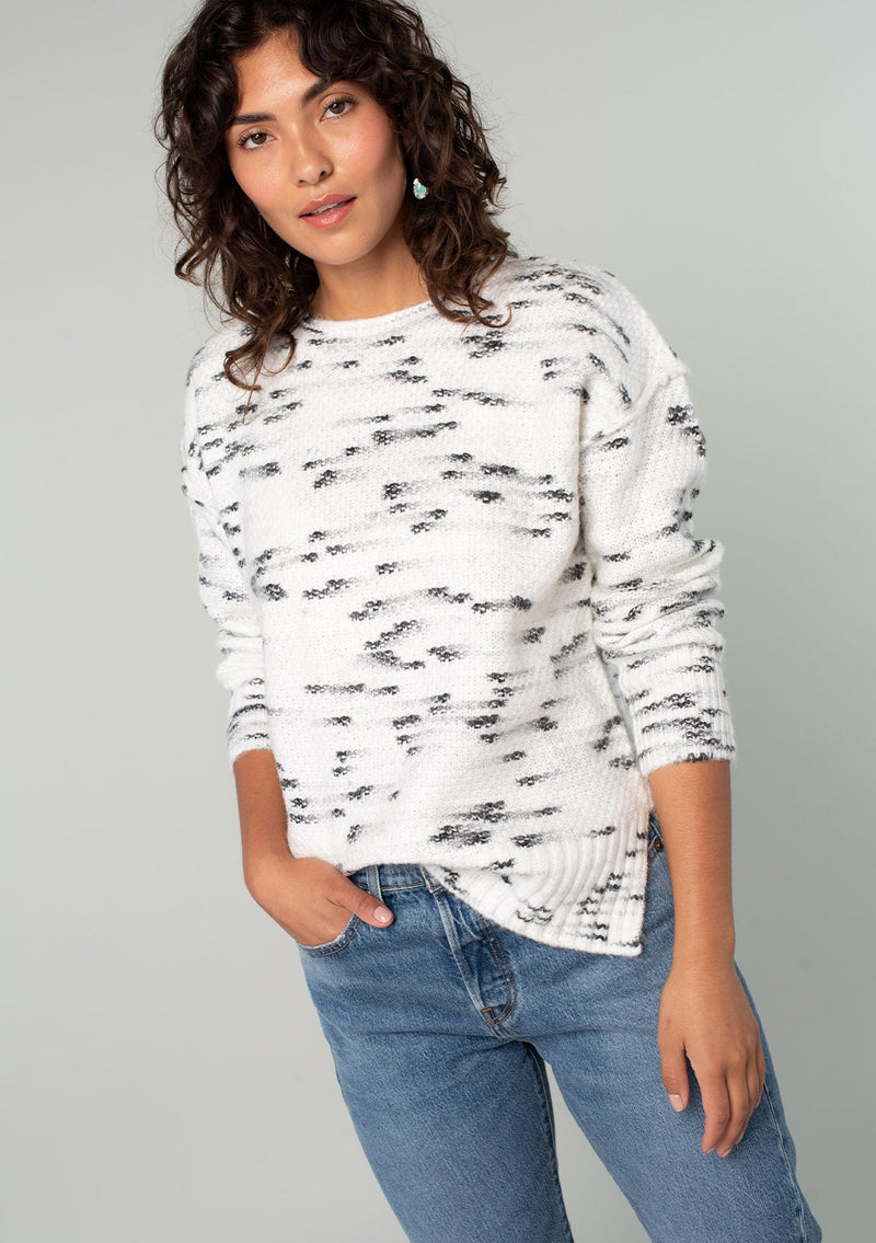 [Color: Ivory/Black] A front facing image of a brunette model wearing a black and white speckled knit pullover sweater. With long sleeves, exposed seam details, and a round neckline. 