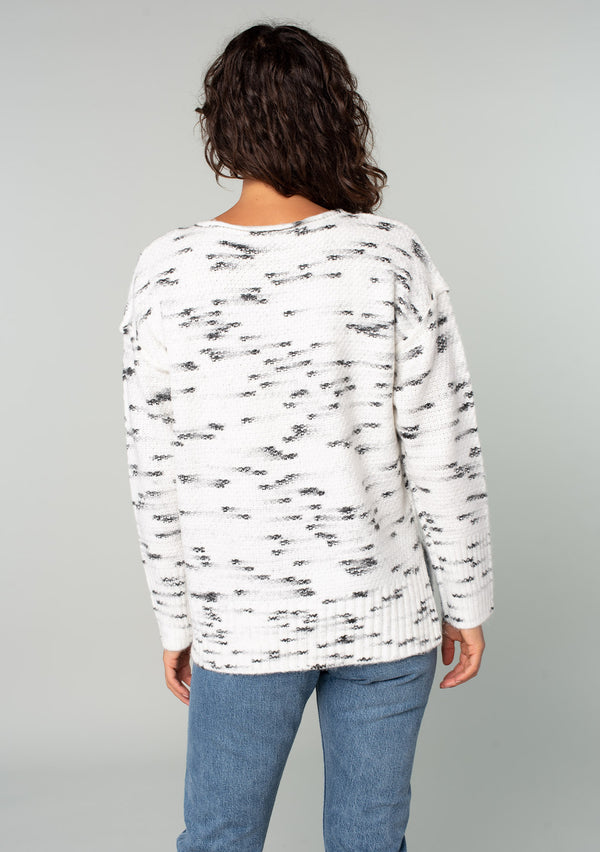 [Color: Ivory/Black] A back facing image of a brunette model wearing a black and white speckled knit pullover sweater. With long sleeves, exposed seam details, and a round neckline. 