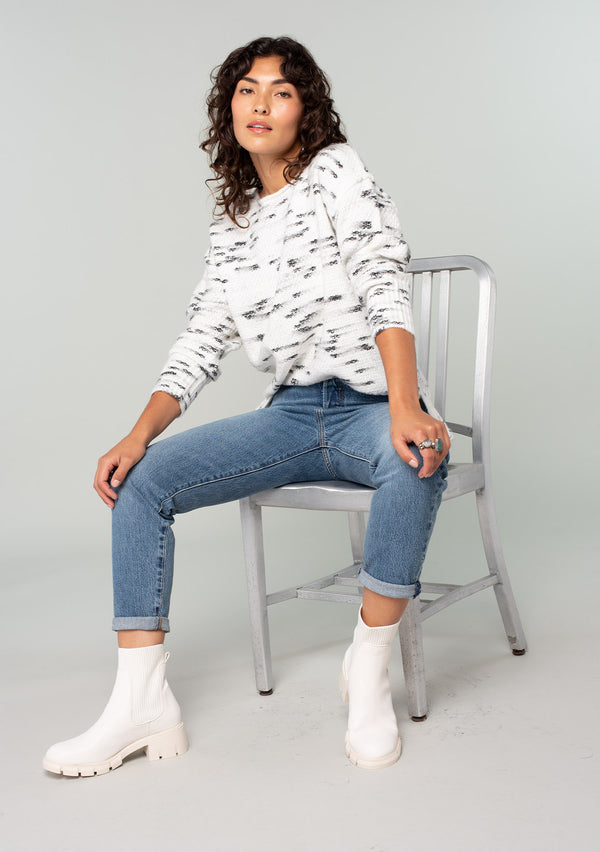 [Color: Ivory/Black] A sitting front facing image of a brunette model wearing a black and white speckled knit pullover sweater. With long sleeves, exposed seam details, and a round neckline. 