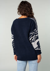 [Color: Navy/Silver] A back facing image of a brunette model wearing a bohemian cotton navy sweater with a silver floral motif. With long sleeves and a crew neckline. 