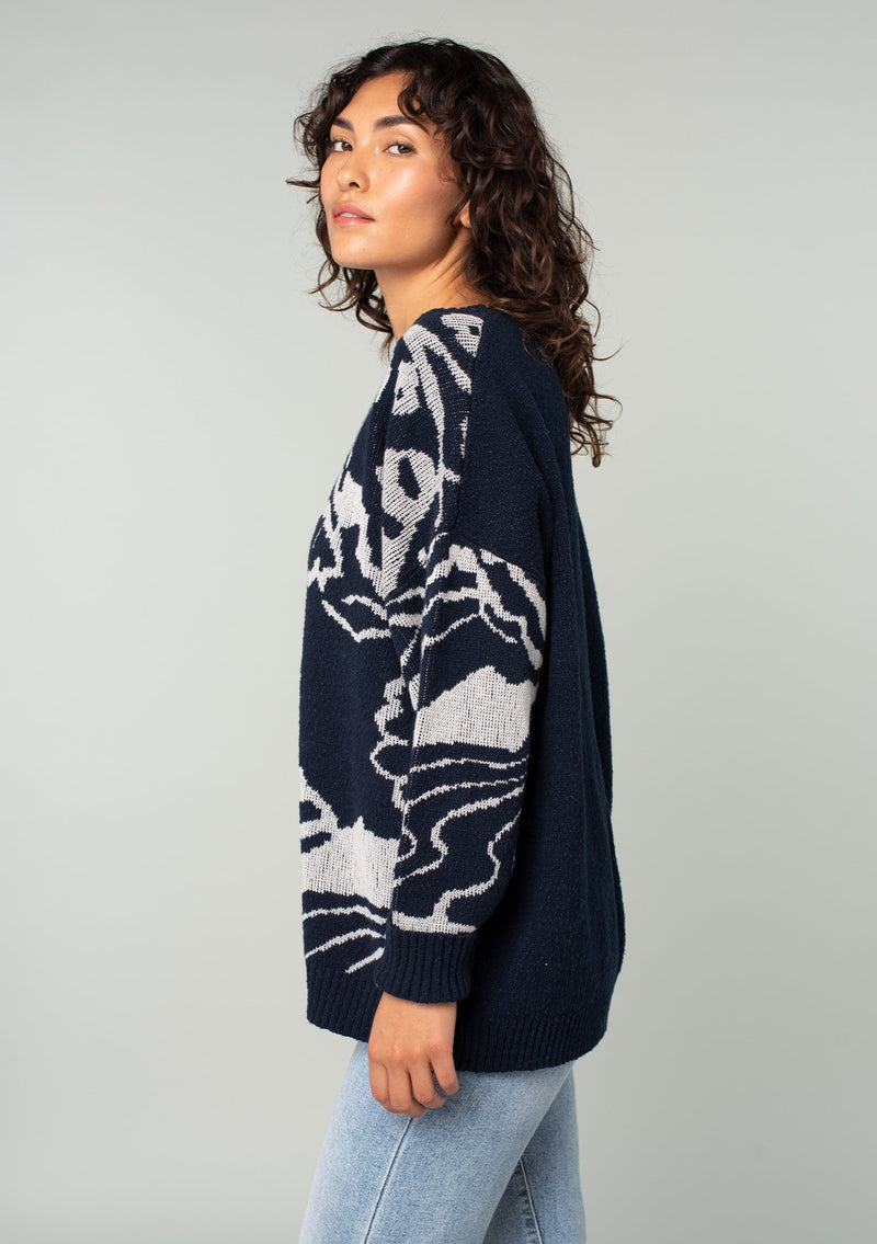 [Color: Navy/Silver] A side facing image of a brunette model wearing a bohemian cotton navy sweater with a silver floral motif. With long sleeves and a crew neckline. 
