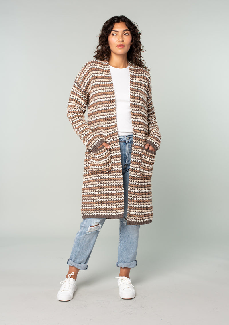 [Color: Mocha Multi] A front facing image of a brunette model wearing a brown multi color stripe chunky knit fall cardigan. A long duster cardigan with an open front, long sleeves, and side pockets with contrast trim. 