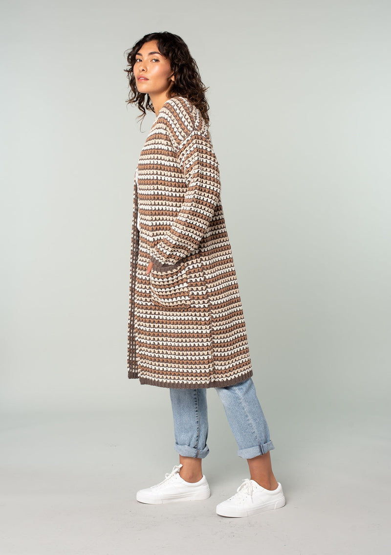 [Color: Mocha Multi] A side facing image of a brunette model wearing a brown multi color stripe chunky knit fall cardigan. A long duster cardigan with an open front, long sleeves, and side pockets with contrast trim. 