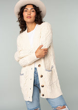 [Color: Natural] A front facing image of a brunette model wearing a cream colored chunky cable knit cardigan. With long sleeves, a v neckline, an oversize button front, and side pockets with contrast blue trim. 