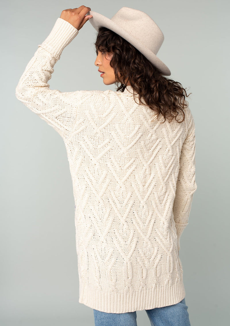 [Color: Natural] A back facing image of a brunette model wearing a cream colored chunky cable knit cardigan. With long sleeves, a v neckline, an oversize button front, and side pockets with contrast blue trim. 