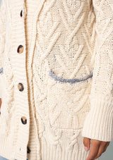 [Color: Natural] A close up front facing image of a brunette model wearing a cream colored chunky cable knit cardigan. With long sleeves, a v neckline, an oversize button front, and side pockets with contrast blue trim.