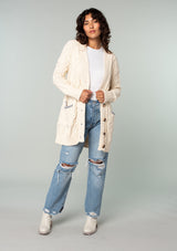 [Color: Natural] A full body front facing image of a brunette model wearing a cream colored chunky cable knit cardigan. With long sleeves, a v neckline, an oversize button front, and side pockets with contrast blue trim. 