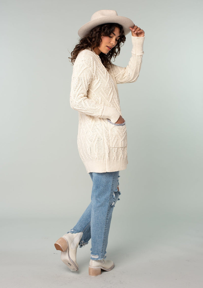 [Color: Natural] A full body back facing image of a brunette model wearing a cream colored chunky cable knit cardigan. With long sleeves, a v neckline, an oversize button front, and side pockets with contrast blue trim. 